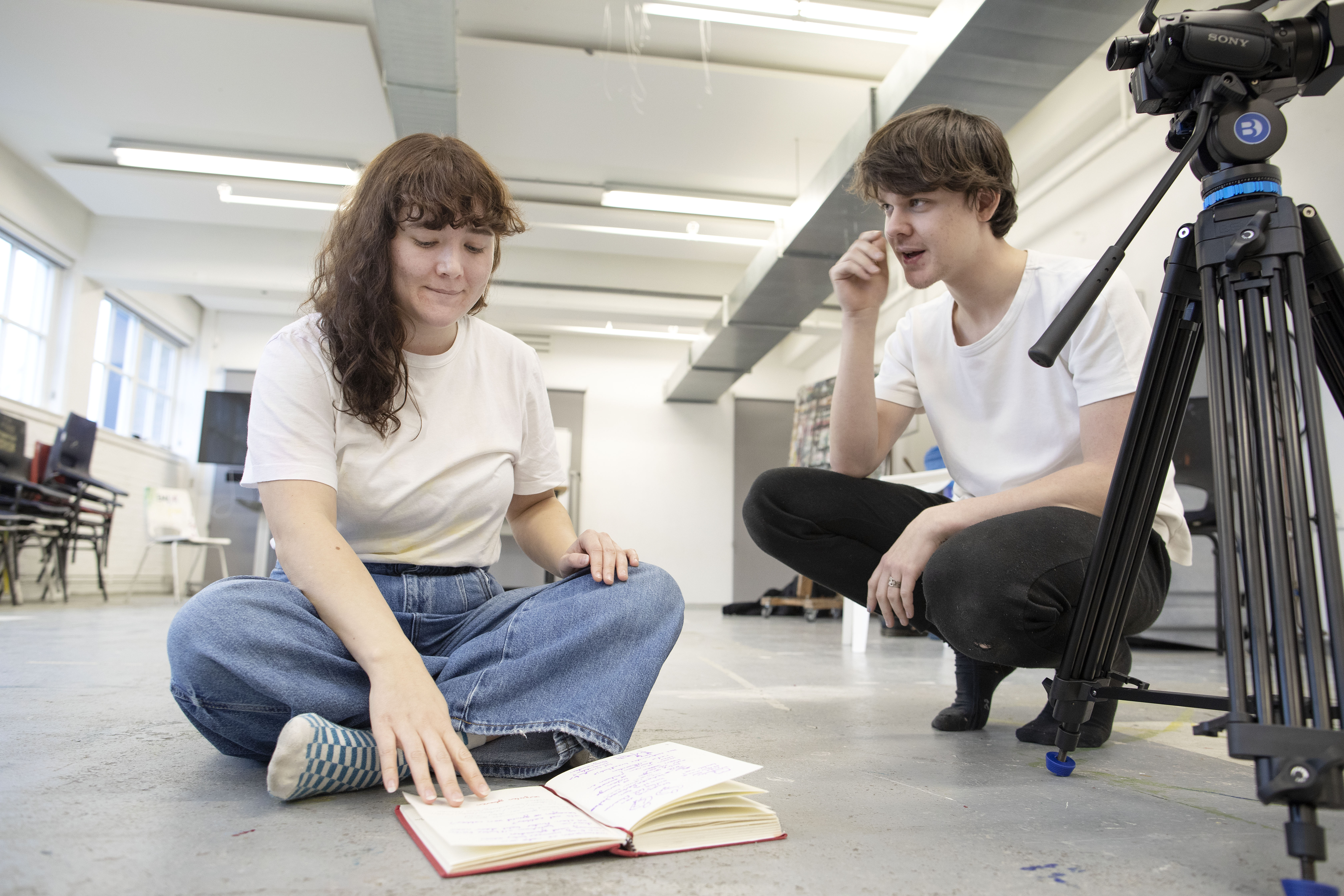 Sanne en partner zitten gehurkt op een grijze vloer in gesprek met een aantekenboekje en filmcamerastatief op de voorgrond